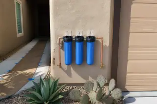 Sediment water filter housing with plumbing beside a garage in a Tempe, AZ desert yard.
