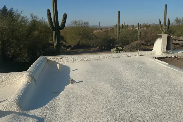 Worker spraying polyurethane foam insulation on a flat roof in Tucson, AZ with desert cacti in the background.