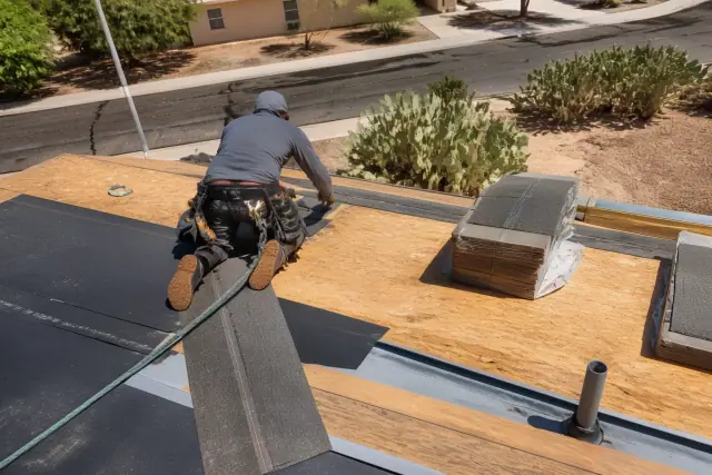 A technician installs shingles on a steep residential roof in Tucson, AZ, with prickly pear cactus in the yard.