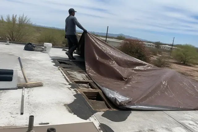 A contractor places a reinforced tarp over a damaged roof during repairs in Flowing Wells, AZ, above desert landscaping.