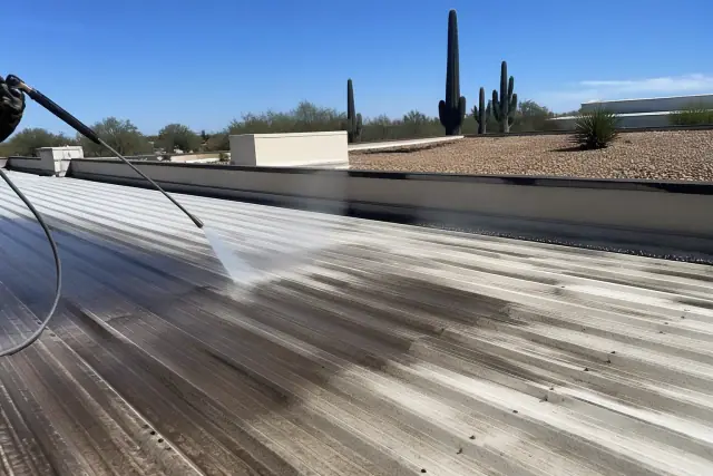 A pressure washer cleans a weathered metal roof in Tucson, AZ, with saguaro cacti and desert landscaping in the background.