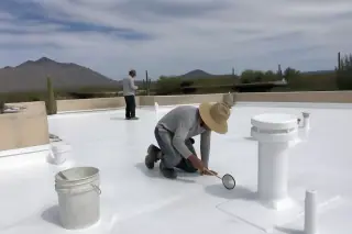Technician inspecting a white reflective roof coating with a mirror near a bucket on a commercial building in Tucson, AZ.