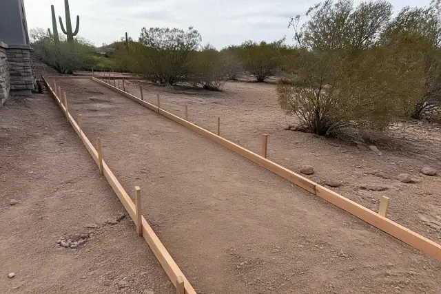 Wooden rails and stakes forming a new sidewalk path in Marana, AZ with desert soil and creosote bushes in the background.