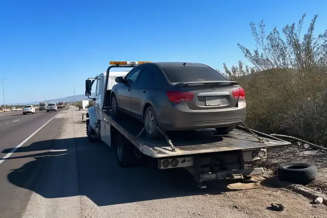 A flatbed tow truck with a disabled sedan on a highway shoulder in Tucson, AZ, surrounded by desert scrub.