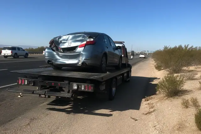 A flatbed tow truck hauling a damaged sedan on a highway shoulder in Drexel Heights, AZ, with desert landscape background.