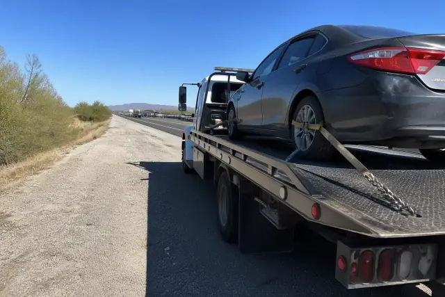 A flatbed truck with a secured sedan on a highway shoulder in Tucson, AZ, surrounded by desert creosote bushes.