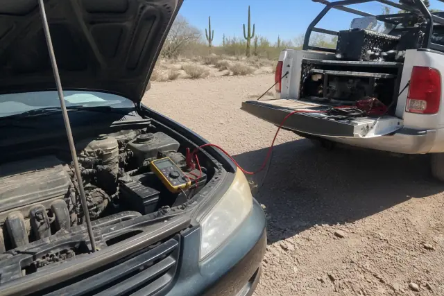 A technician performs a battery load test with a multimeter on a sedan parked on gravel in Tucson, AZ.