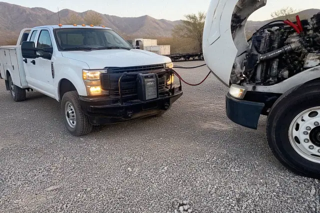 Diesel truck with hood raised and jumper cables attached at a gravel fleet yard in Tucson, AZ with mountain views.