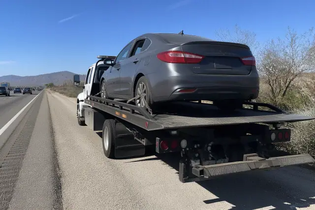 A flatbed tow truck with a disabled sedan on the highway shoulder in Tanque Verde, AZ, surrounded by Sonoran desert scrub.