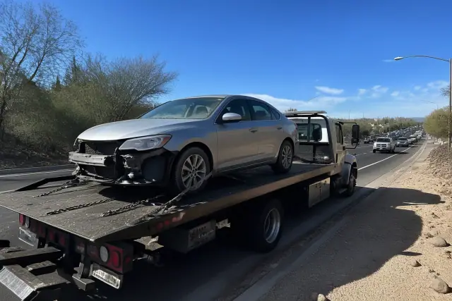 Flatbed tow truck with a secured sedan on the side of a road in Tanque Verde, AZ, with desert soil and creosote bushes.