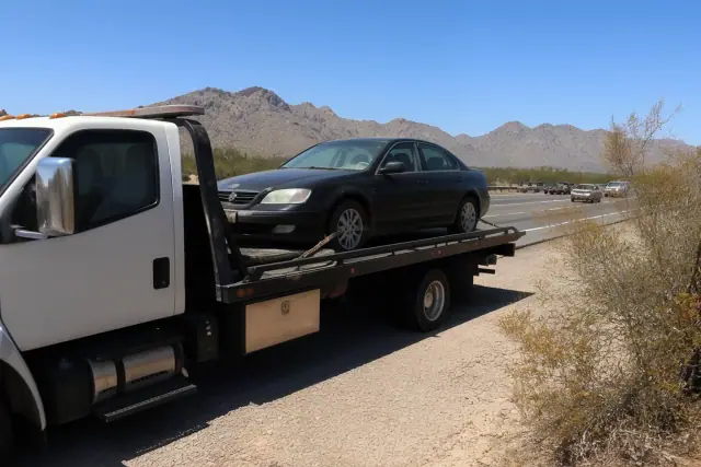 A flatbed tow truck with a disabled sedan secured on a desert road in Tucson, AZ, with mountains in the background.