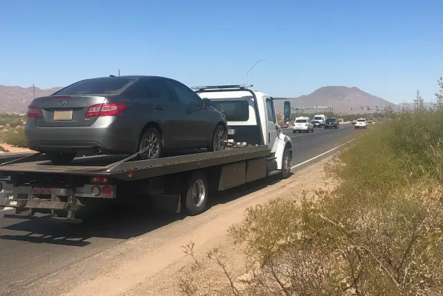 A flatbed tow truck with a disabled sedan on the back parked on a roadside in Tucson, AZ, with desert mountains.