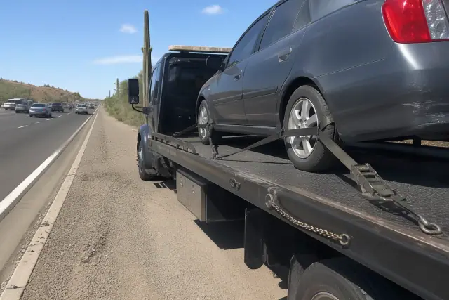Flatbed tow truck securing a sedan with straps on a highway shoulder in a Tucson, AZ desert landscape.