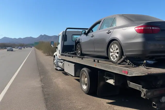 Flatbed tow truck with a disabled sedan on a highway shoulder in Tucson, AZ, with desert mountains in the background.