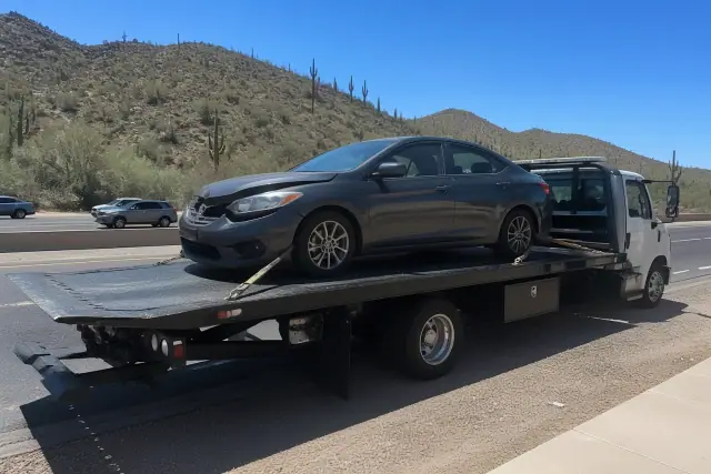 A flatbed tow truck carrying a disabled sedan on a busy road in Tucson, AZ, with desert hills in the background.