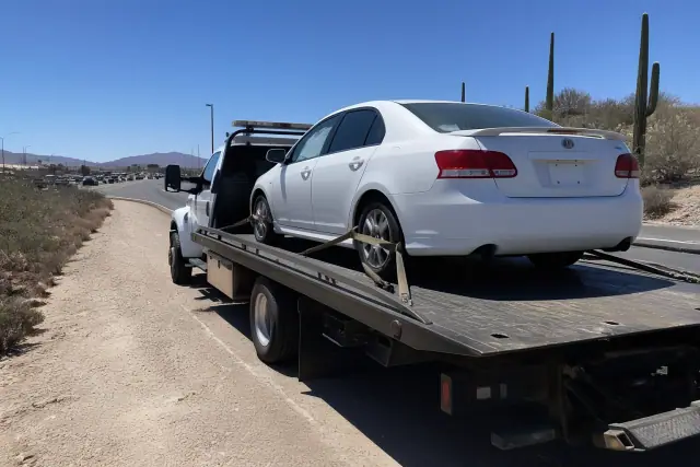 A flatbed tow truck with a disabled sedan secured on a highway shoulder in Tucson, AZ, surrounded by desert landscape.