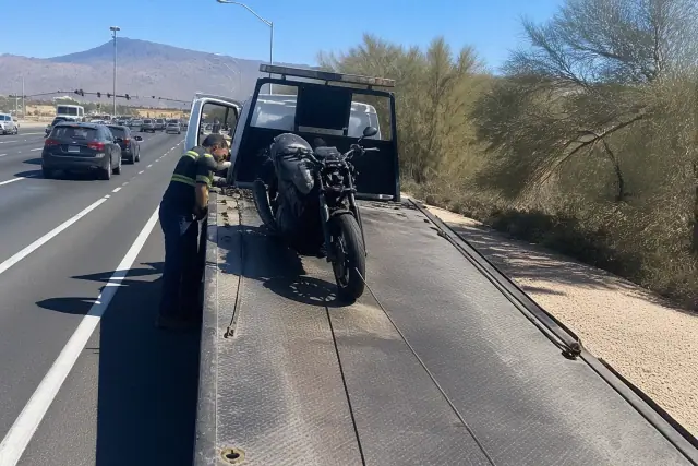 A damaged motorcycle being winched onto a flatbed truck on a roadside in Tucson, AZ, with desert mountains in the background.