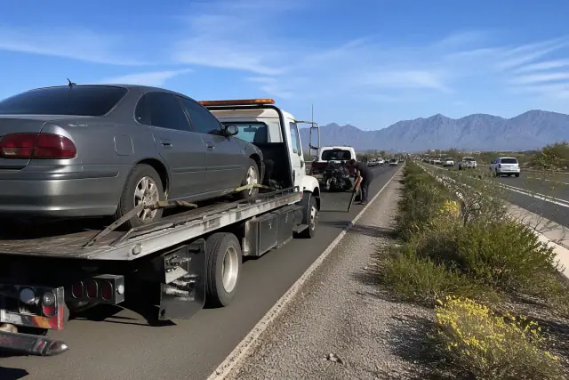 Flatbed truck loading a damaged motorcycle and a disabled sedan on a highway shoulder in Tucson, AZ.