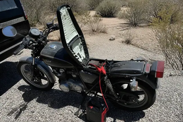 A motorcycle with an open seat and a portable jump pack connected to the battery on gravel in Tucson, AZ.