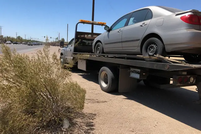 Flatbed truck hauling a disabled sedan on a busy roadside in Flowing Wells, AZ, surrounded by desert brush.