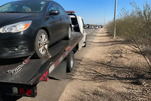 A flatbed tow truck with a secured sedan on a roadside in Tucson, AZ, with desert scenery and safety chains visible.