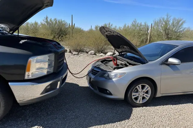 A jump-start service vehicle connected by cables to a dead sedan in a gravel lot with saguaro cacti in Tucson, AZ.