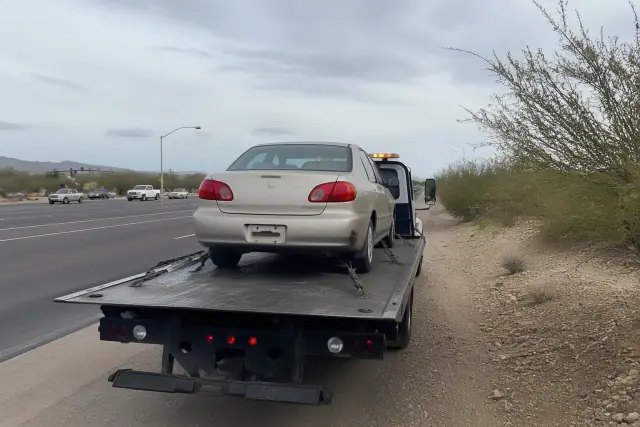 A disabled sedan secured with four tie-down straps on a flatbed truck on a road in Tucson, AZ.