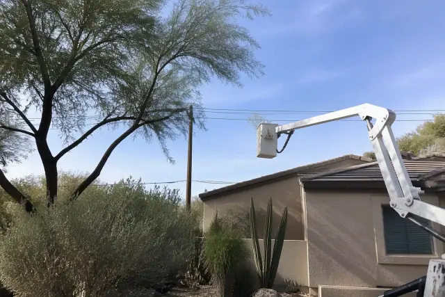 Articulated boom lift trimming tree branches away from a stucco house roofline in Tucson, AZ.